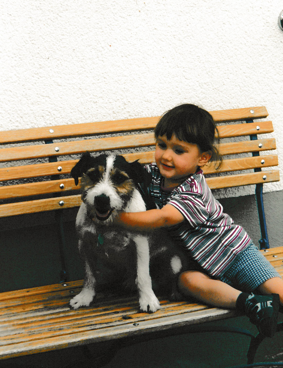 Kleinkind mit gestreiftem Shirt umarmt einen Jack Russel Terrier auf einer Holzbank – liebevolle, nostalgische Momentaufnahme.